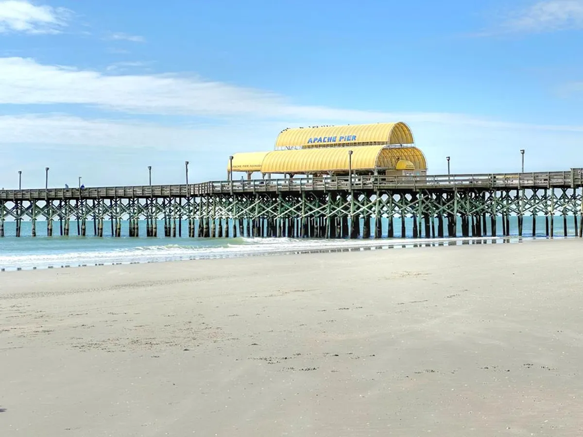 This image features the iconic Apache Pier stretching out into the Atlantic Ocean, with soft sandy beaches and gentle waves under a partly cloudy sky. People can be seen walking along the pier and enjoying the relaxing beach atmosphere.