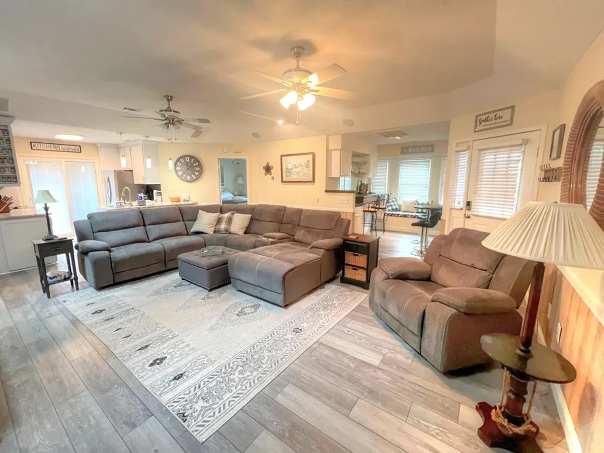 A cozy living room scene featuring a large sectional sofa atop an area rug, a floor lamp, and a dining area visible in the background. The space exudes an inviting, homey feel, perfect for relaxing after a day at the beach or enjoying board games with family.