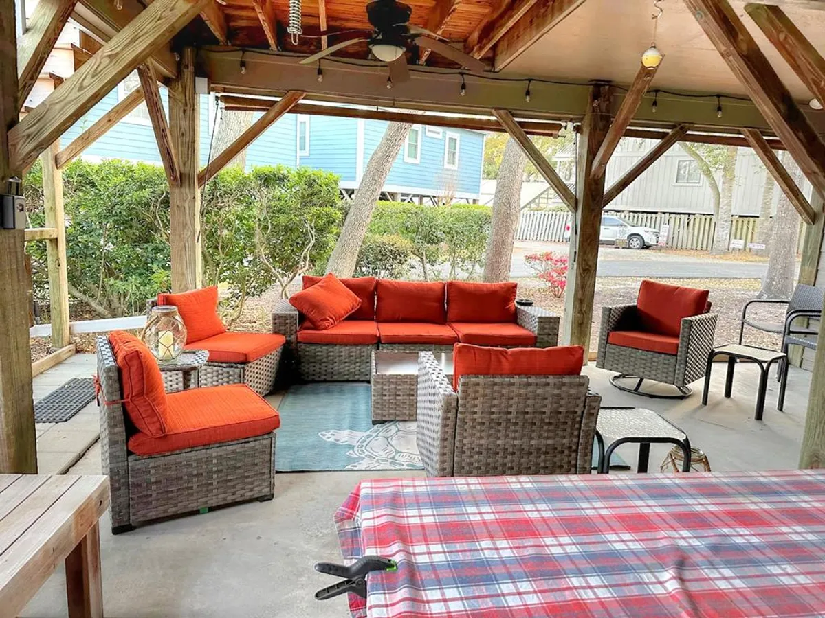 An inviting outdoor patio area with cozy woven furniture, bright orange cushions, and a dining table set under the open sky. The space encourages social gatherings, outdoor dining, and relaxing after a day at the beach.