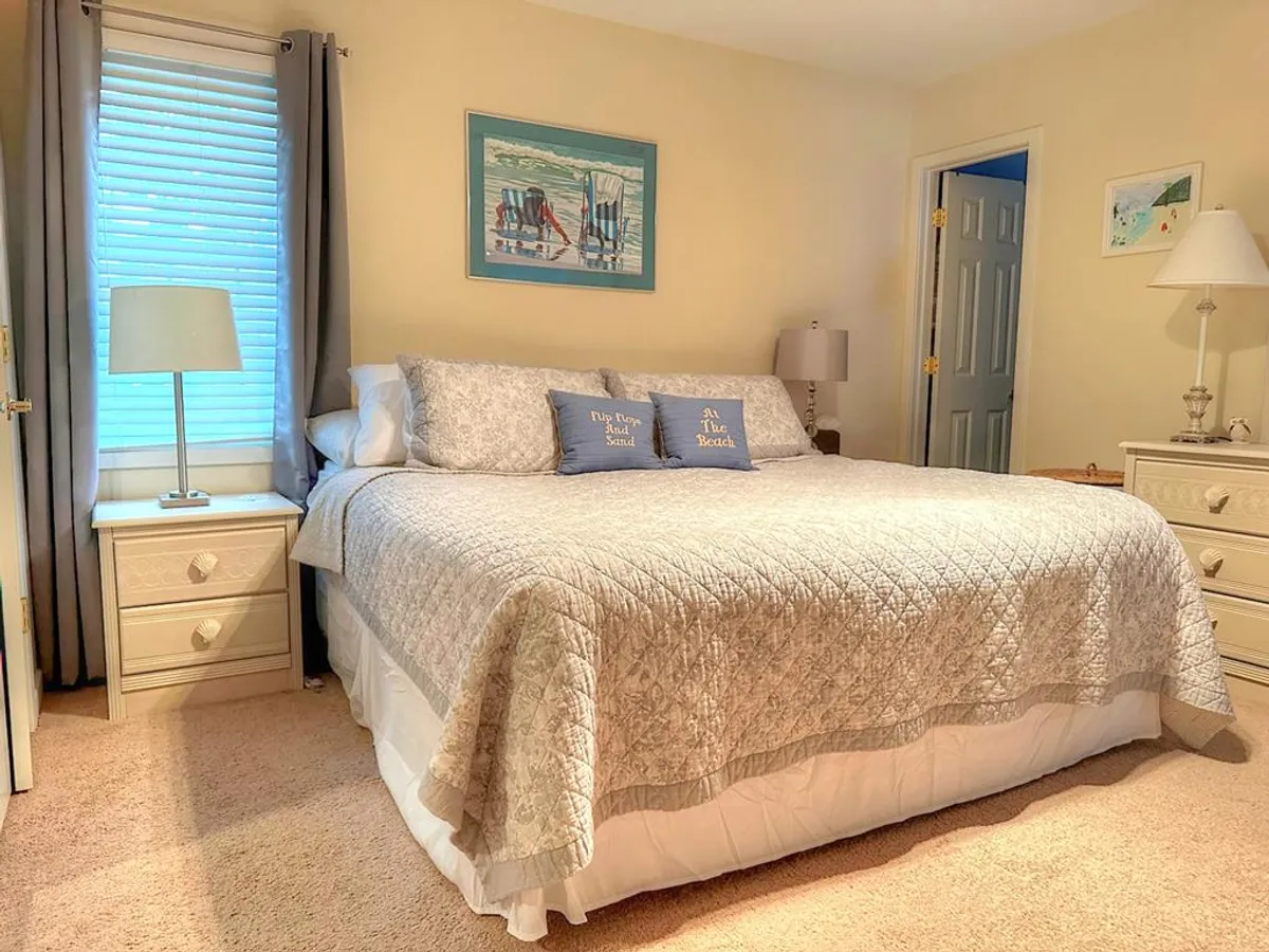 A serene bedroom with a large bed adorned with decorative pillows and cozy linens. Nightstands with lamps flank the bed, and framed artwork creates a calming atmosphere ideal for rest and relaxation.