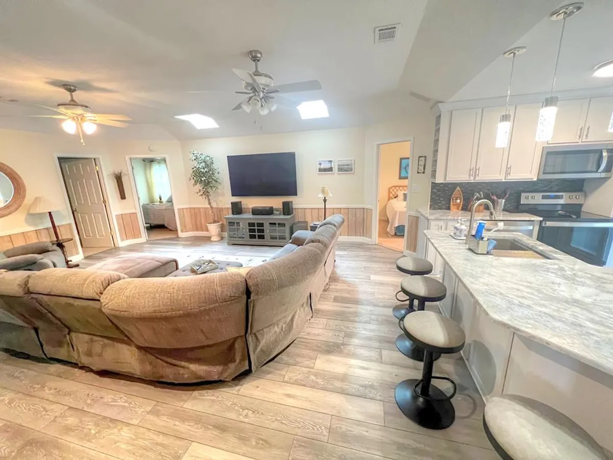 This image captures an open-concept living area with a comfortable sectional sofa facing a large flat-screen TV. The kitchen island with seating and a dining area are visible in the background, showcasing the seamless flow between spaces. Natural light brightens the room, creating a welcoming and social atmosphere.