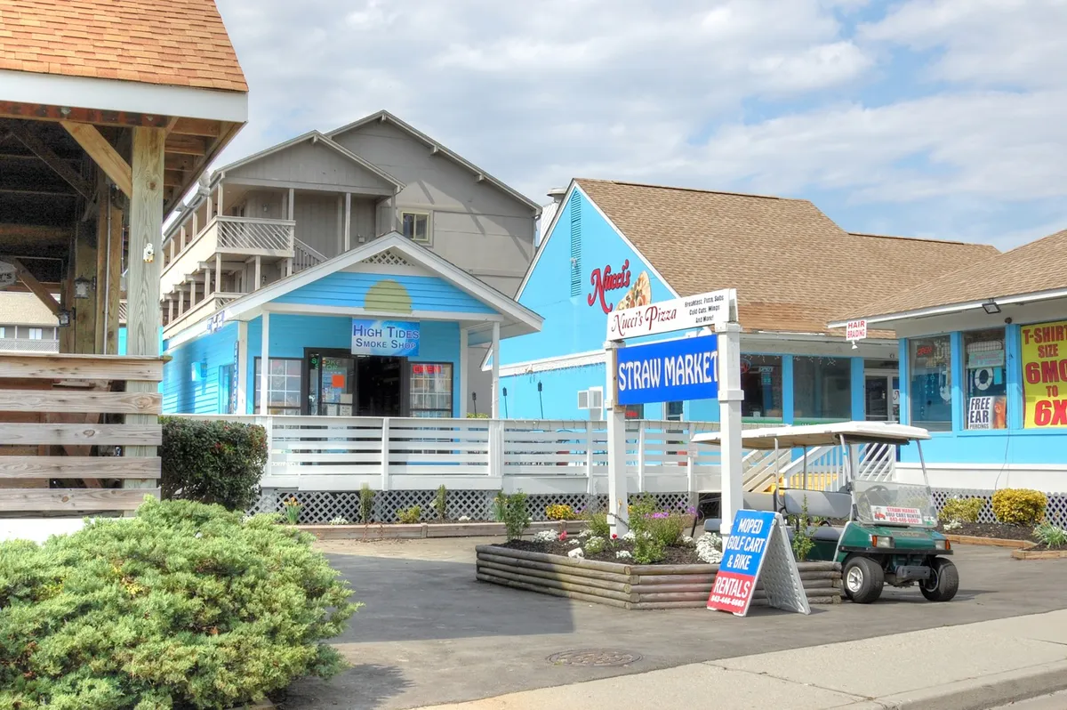 This cheerful street scene features colorful storefronts, the Straw Market sign, a golf cart ready for exploration, and people enjoying the sunshine as they visit local shops and eateries. The overall mood is laid-back, vibrant, and perfect for casual group outings or discovering new favorites together.