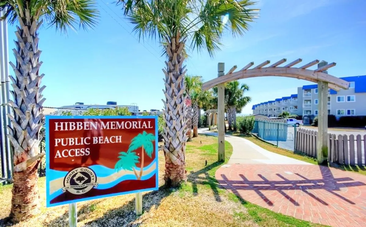 An inviting exterior scene showing the Hibben Memorial Public Beach Access sign, flanked by palm trees and an arched wooden walkway leading toward the sand. The atmosphere is relaxed and welcoming, evoking the proximity and openness to outdoor experiences.