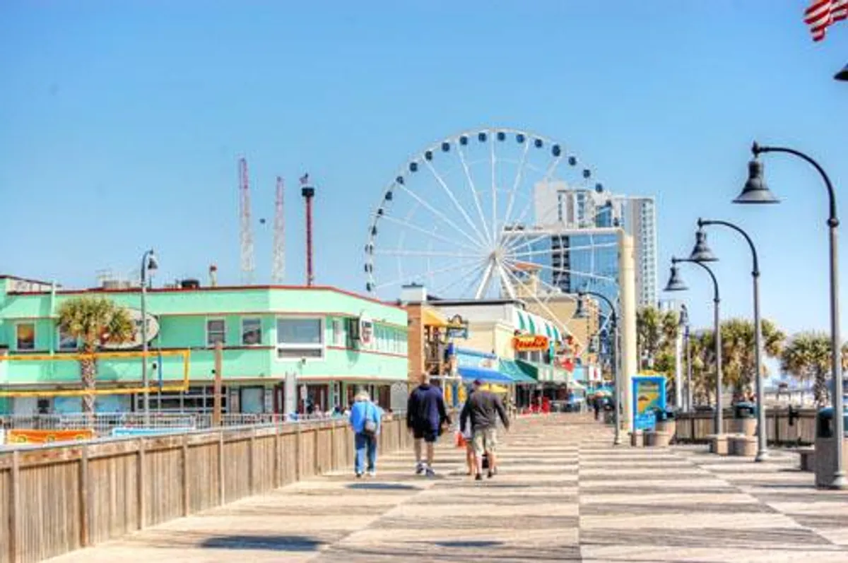 This image captures the iconic Myrtle Beach boardwalk scene, featuring the vibrant ferris wheel, colorful buildings, and groups of people strolling and enjoying a lively atmosphere along the waterfront. The presence of crowds, entertainment venues, and coastal views embodies the bustling energy of Myrtle Beach attractions just steps from the property.