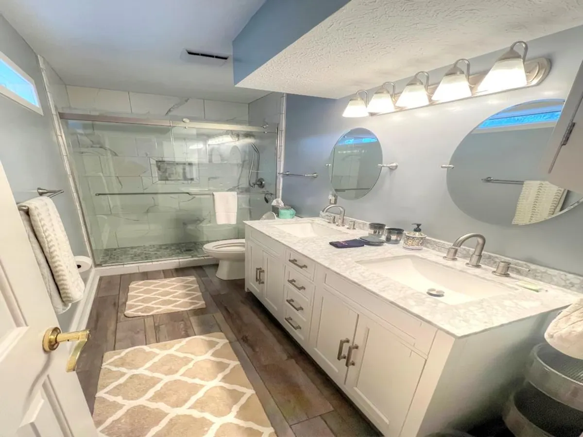 A bright bathroom featuring a double vanity with sinks, a glass shower enclosure, large mirrors, and a decorative rug. The layout and fixtures suggest comfort and convenience for guests seeking privacy or personal space.