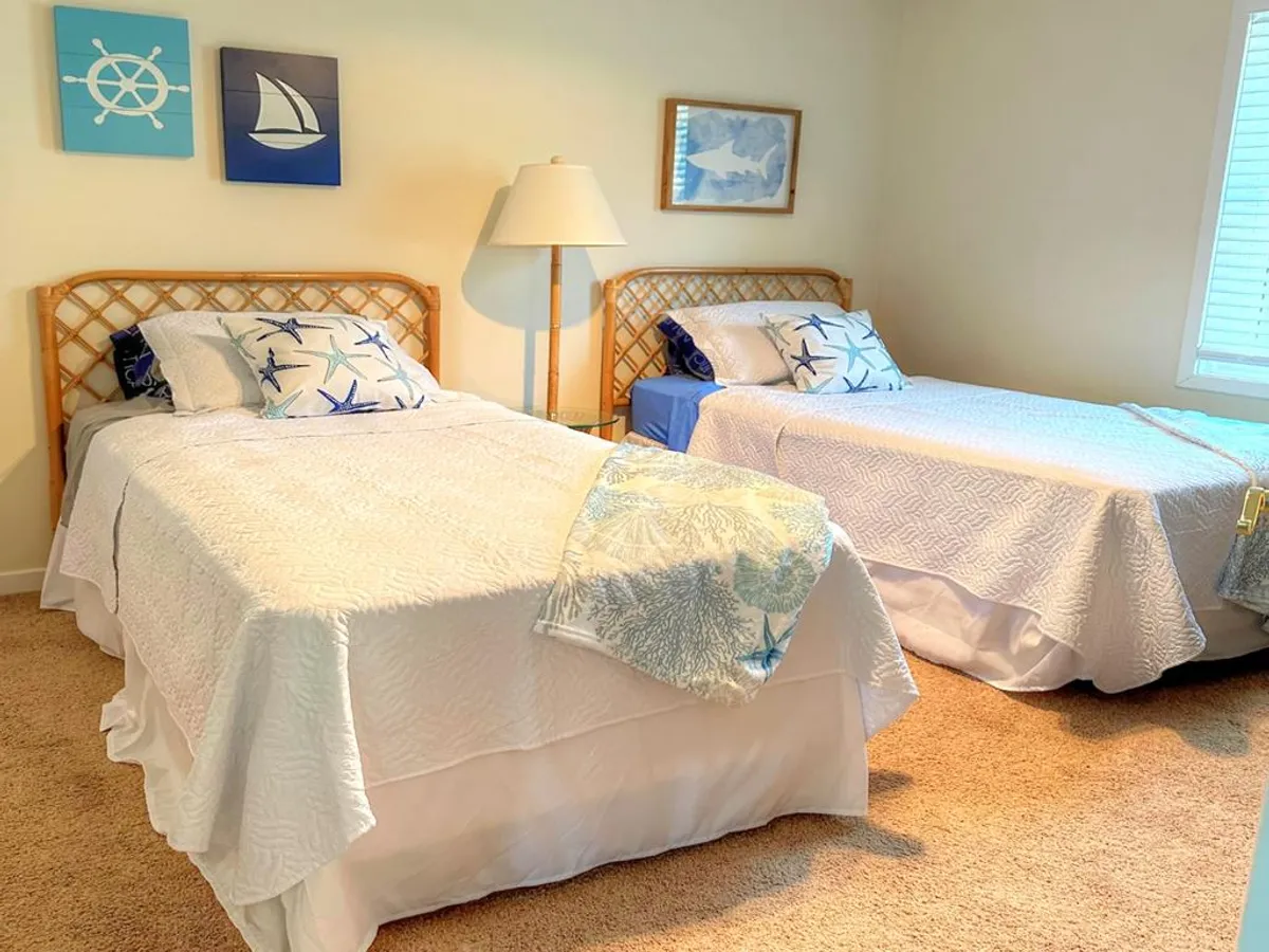 A cheerful twin bedroom with two twin beds, colorful beach-themed artwork, decorative pillows, and a lamp. The space is bright and inviting, perfect for kids or guests seeking a relaxed sleeping arrangement.