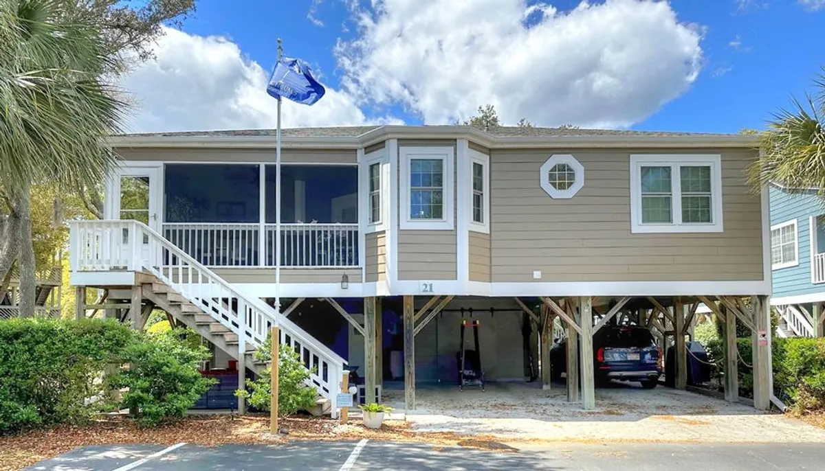 The image showcases the stilted exterior of a charming, two-story cottage in Myrtle Beach. A welcoming front porch with comfortable seating invites guests to relax, while lush landscaping and a distinctive blue flag add coastal flair. The scene hints at leisurely mornings, vibrant sunsets, and space for family gatherings or barbecues.