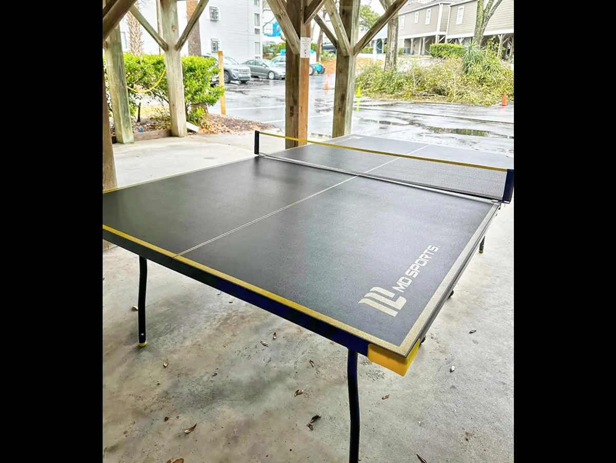 A vibrant outdoor scene featuring a ping pong table set beneath the cottage, surrounded by wooden support beams and lush greenery. The area is clearly designed for casual gatherings and friendly competition, inviting guests to relax and play.