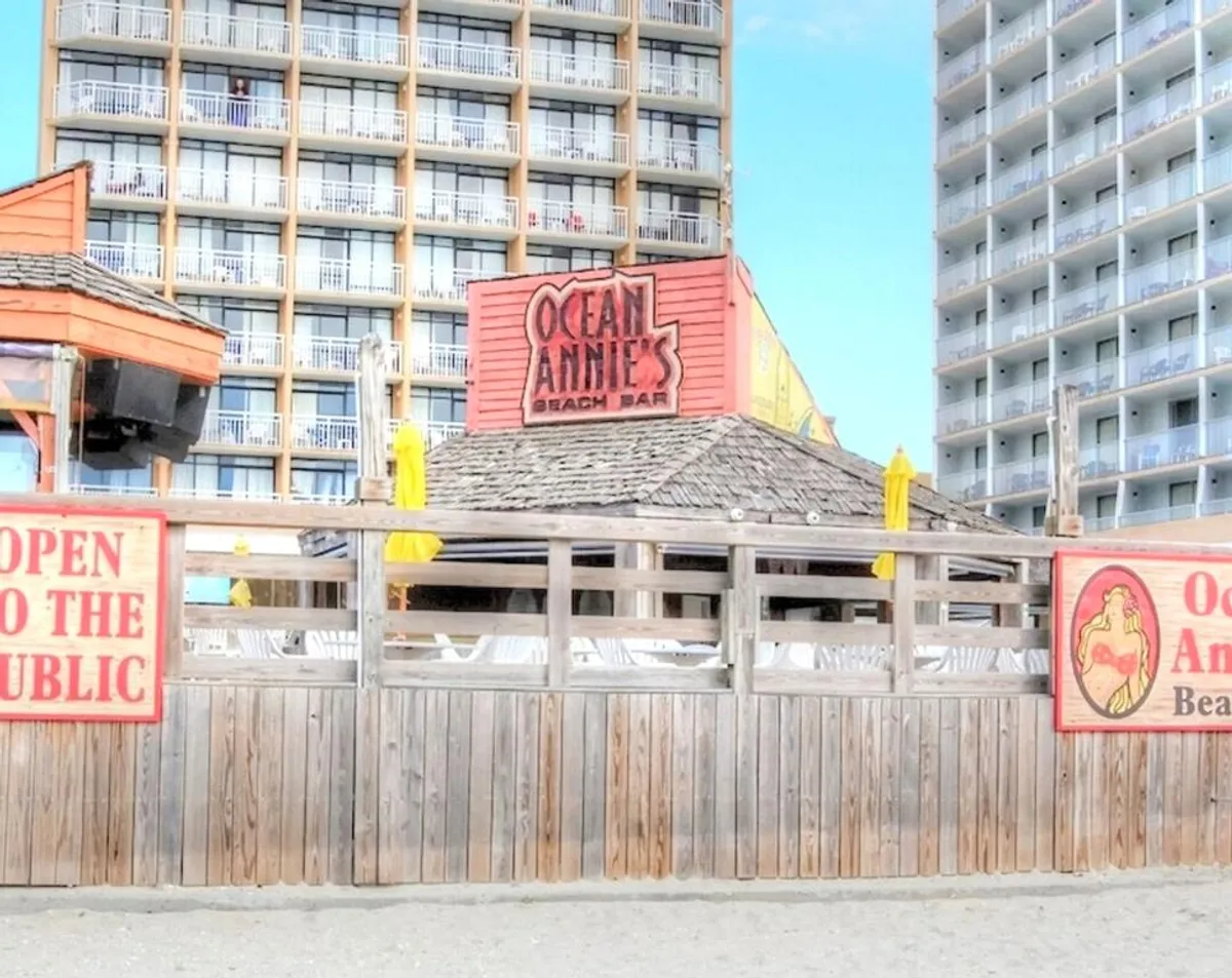 The photo spotlights the festive exterior of Ocean Annie's Beach Bar, with its vibrant yellow umbrellas, wooden fence, and groups dining and socializing in a lively beachside setting. The sunset lighting, visible in the background, adds a relaxed, fun-filled vibe, ideal for gatherings and outdoor entertainment.