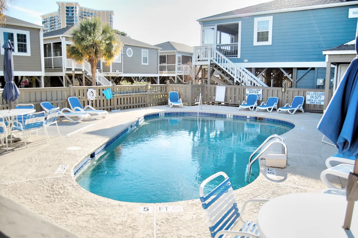 A sparkling community pool bordered by lounge chairs, palm trees, and neighboring cottages. The area is inviting and designed for both relaxation and socializing, with ample space for sunbathing and swimming.